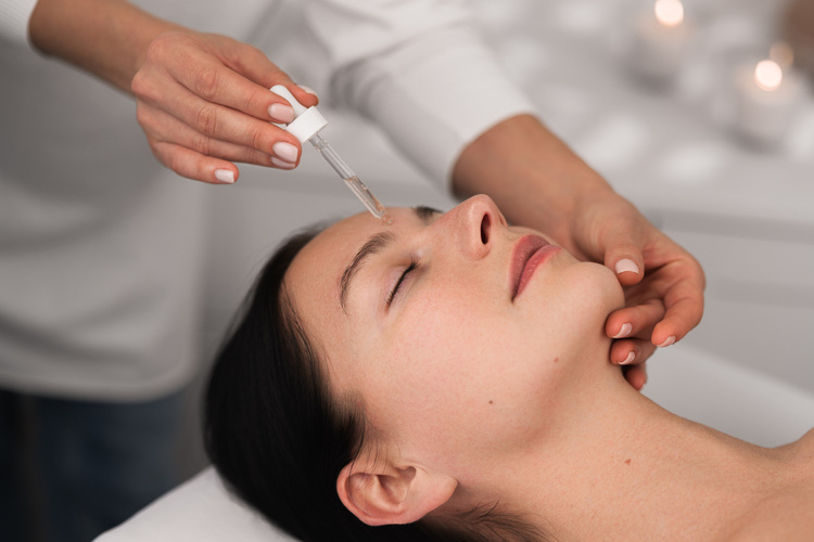 High angle of crop anonymous beautician applying serum with dropper on face of calm young female client, lying on table with closed eyes during skincare procedure in salon